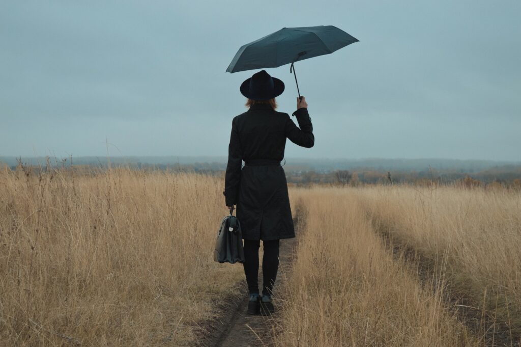 Woman on a journey walking through a field carrying an open black umbrella and a satchel.